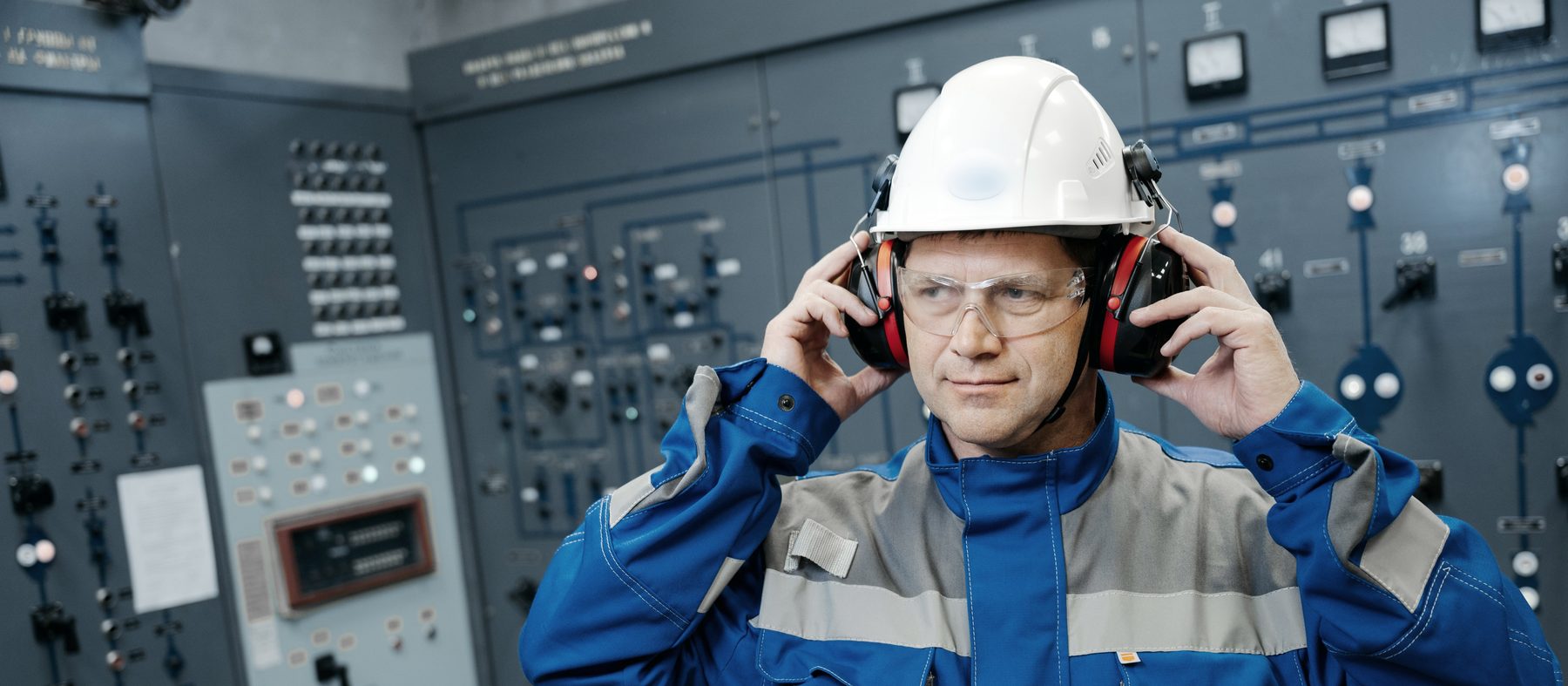 Trabalhador ajustando protetor auricular, com capacete e óculos de proteção, diante de painéis elétricos